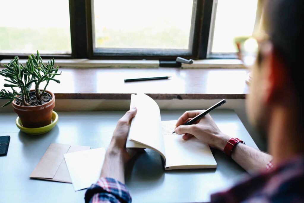 Man in glasses writing in a book at a window
