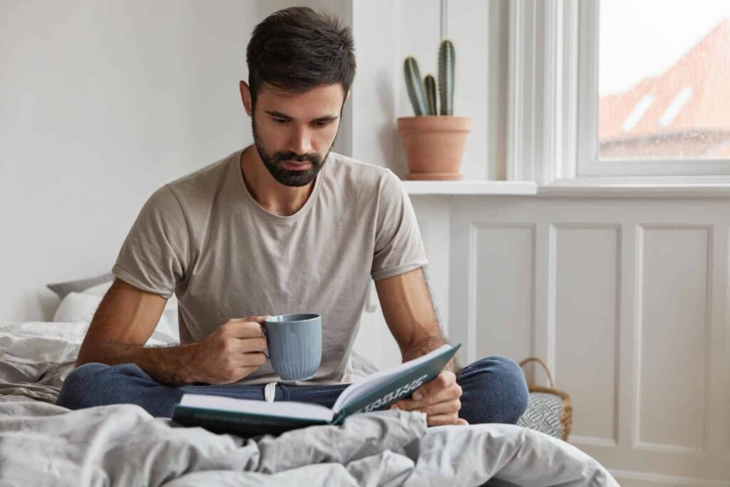 Indoor shot of handsome young hipster with thick beard, dressed casually, sits on bed, busy with reading, drinks fresh aromatic coffee, poses in bedroom. People, hobby and lifestyle concept.